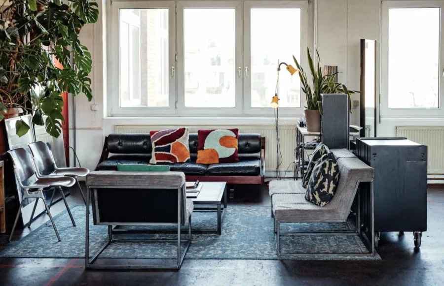 Lounge-Bereich mit Tageslicht im ikonic STUDIO Berlin: Schwarzes Ledersofa, bunte Kissen, Design-Sessel auf einem blauen Teppich und große Fensterfront für Events und Meetings.